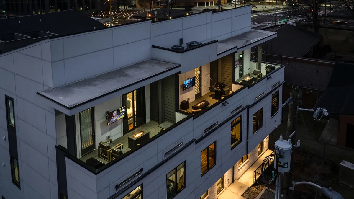 A modern rooftop terrace featuring comfortable outdoor seating, illuminated windows, and television screens. The atmosphere is sophisticated and inviting, with city lights visible in the background, suggesting a perfect space for evening gatherings or rooftop parties.