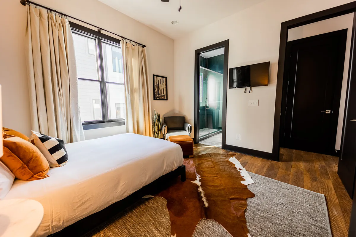 A cozy bedroom featuring a bed with decorative pillows, a large window with natural light, stylish artwork, a shower area with a glass door, and a wall-mounted television. The space seamlessly blends sleeping and bathing amenities, offering both convenience and comfort.