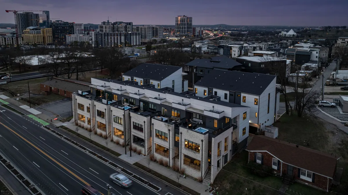 A row of modern townhouses lines a tree-shaded urban street, with the Nashville city skyline visible in the background. The scene exudes an inviting yet sophisticated mood, suggesting a vibrant, walkable neighborhood perfect for families to explore together. Balconies and outdoor spaces hint at communal living and city engagement.