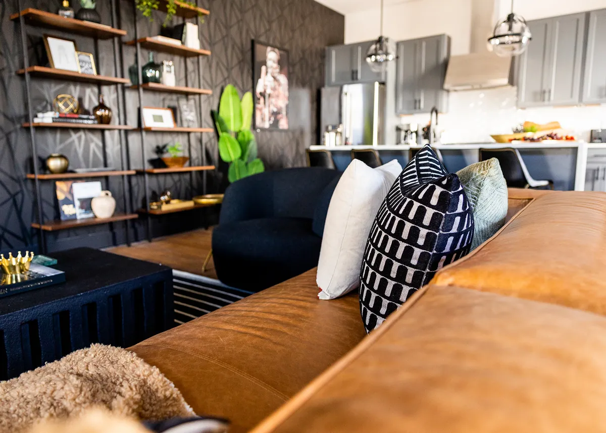 A chic living space with a leather sofa, black accent chair, decorative shelves, green plant, and the kitchen visible in the background. The design is both comfortable and stylish, ideal for entertaining guests.