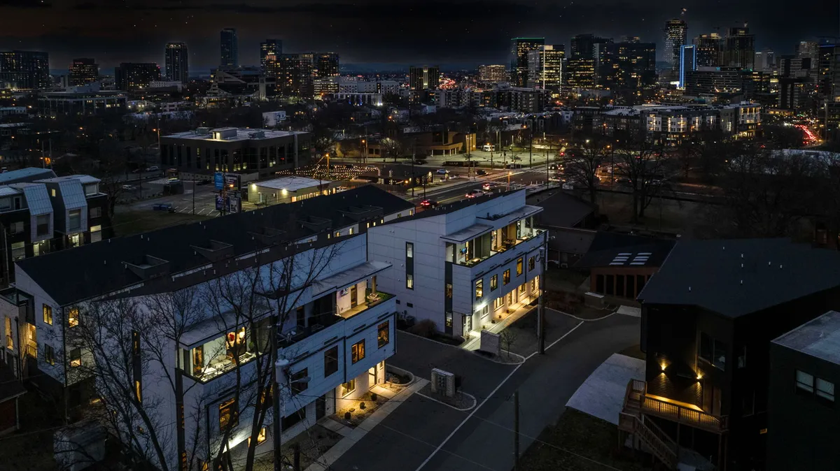 A contemporary exterior shot of modern townhouses set against a city skyline at dusk. Streetlights glow, cars line the road, and the atmosphere is both vibrant and sophisticated, suggesting an ideal spot for group gatherings before hitting the town.