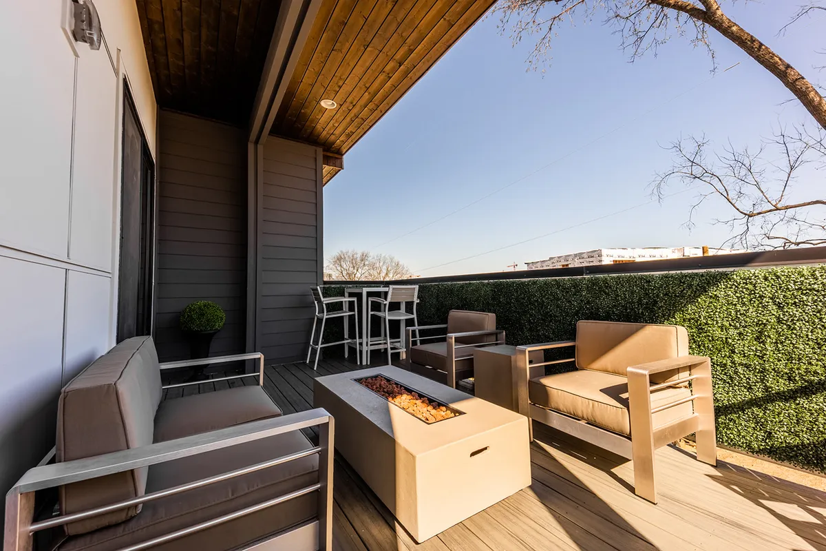 A rooftop deck with a spacious seating area arranged around a glowing fire pit, surrounded by lush greenery and offering an open view of the Nashville skyline. The setup suggests a comfortable environment ideal for both lively group celebrations and relaxed evenings.