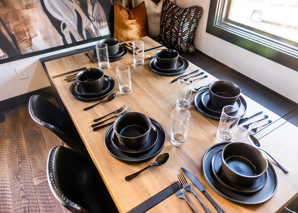 A tastefully set dining table with black plates, glasses, and cutlery, surrounded by decorative pillows, evoking a mood of warmth, celebration, and togetherness.