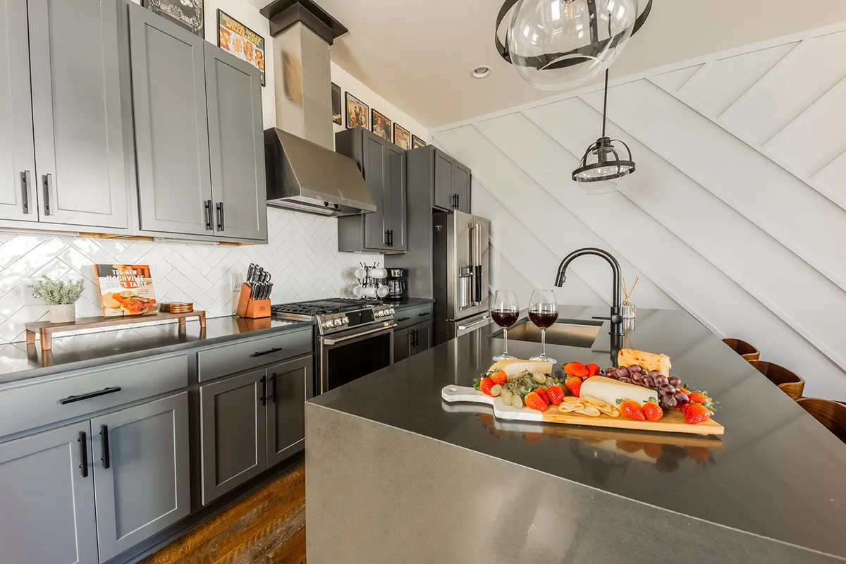 This image features a contemporary kitchen with a central island topped by a charcuterie board and wine glasses. Modern cabinetry and gleaming stainless steel appliances anchor the space, complemented by stylish accents and warm lighting.