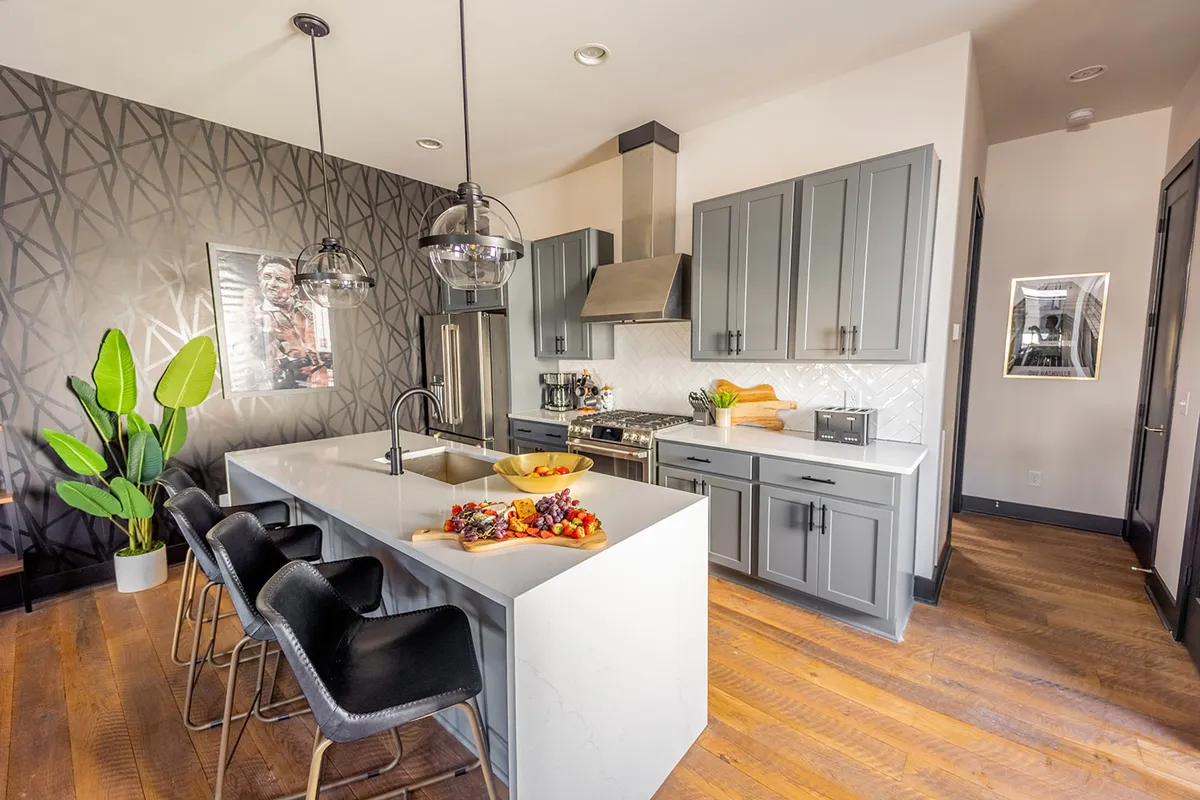A bright, lively kitchen scene with a spacious island, colorful fruit bowl, wall art, and a potted plant. The kitchen is designed for both culinary activity and social interaction, inviting guests to participate in cooking or gather around the island.