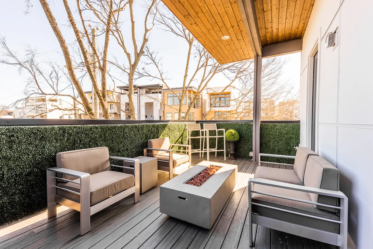 A sleek, contemporary rooftop deck featuring a modern seating area, a cozy fire pit, green hedge accents, and an outdoor dining table. The space is set for socializing, dining al fresco, and enjoying sunset views with friends or family.