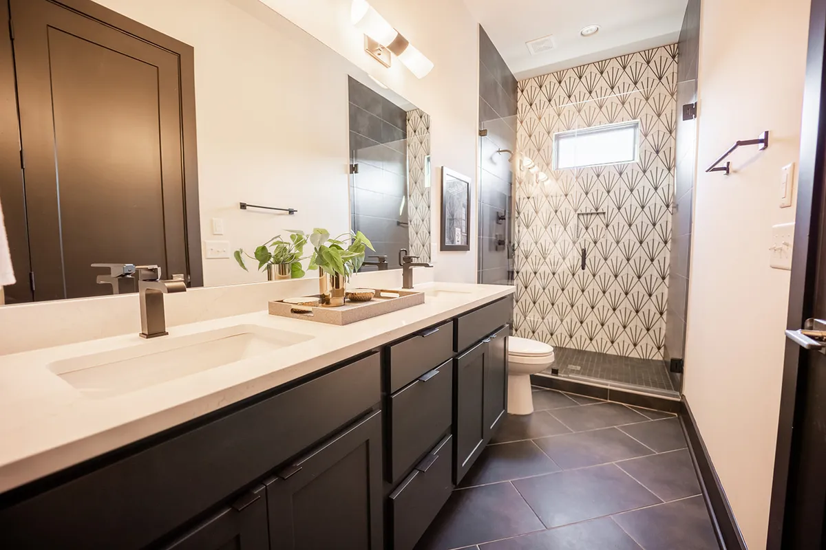 A modern bathroom with double sinks, geometric tile shower wall, black cabinetry, and a small potted plant. The space feels fresh and inviting, reflecting spa-inspired luxury and comfort ideal for unwinding at the end of the day.